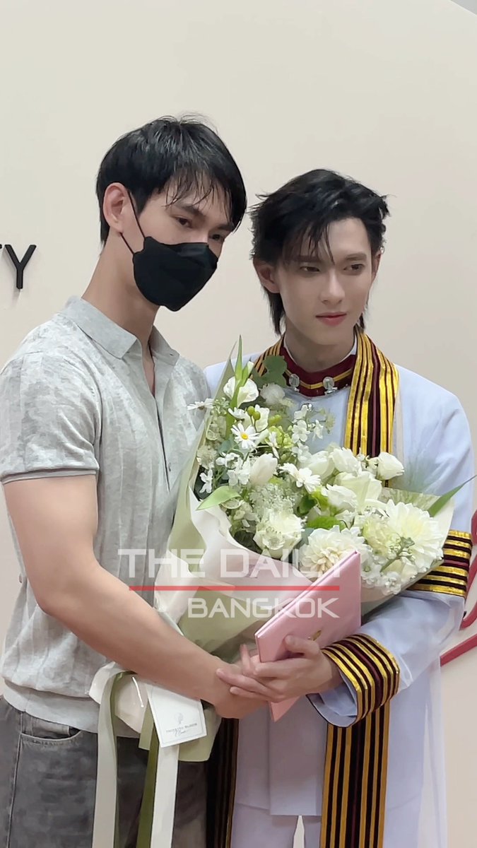 Two young men pose together, with the one on the left wearing a mask and gray shirt while holding a bouquet of white flowers alongside the graduate on the right, who wears a white graduation gown with a striped stole; the image is watermarked "THE DAILY BANGKOK" and accompanies a post celebrating Pooh Krittin's graduation in Robotics & AI Engineering.