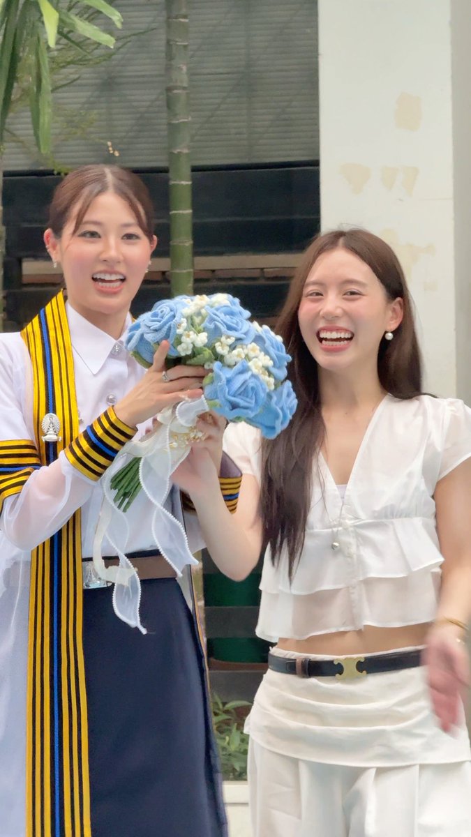 Two smiling young women pose together; the one on the left wears a white shirt and yellow-and-blue striped sash, while the one on the right wears a white top and holds a bouquet of blue roses.