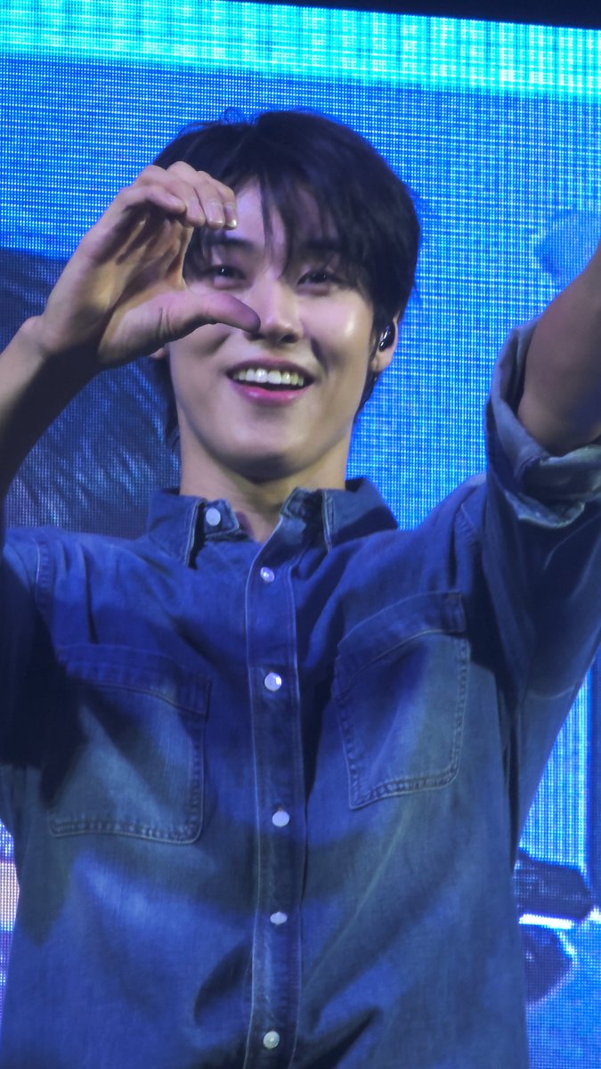 A young man with dark hair in a denim shirt smiles while making a heart gesture with his hands against a bright blue LED screen backdrop.