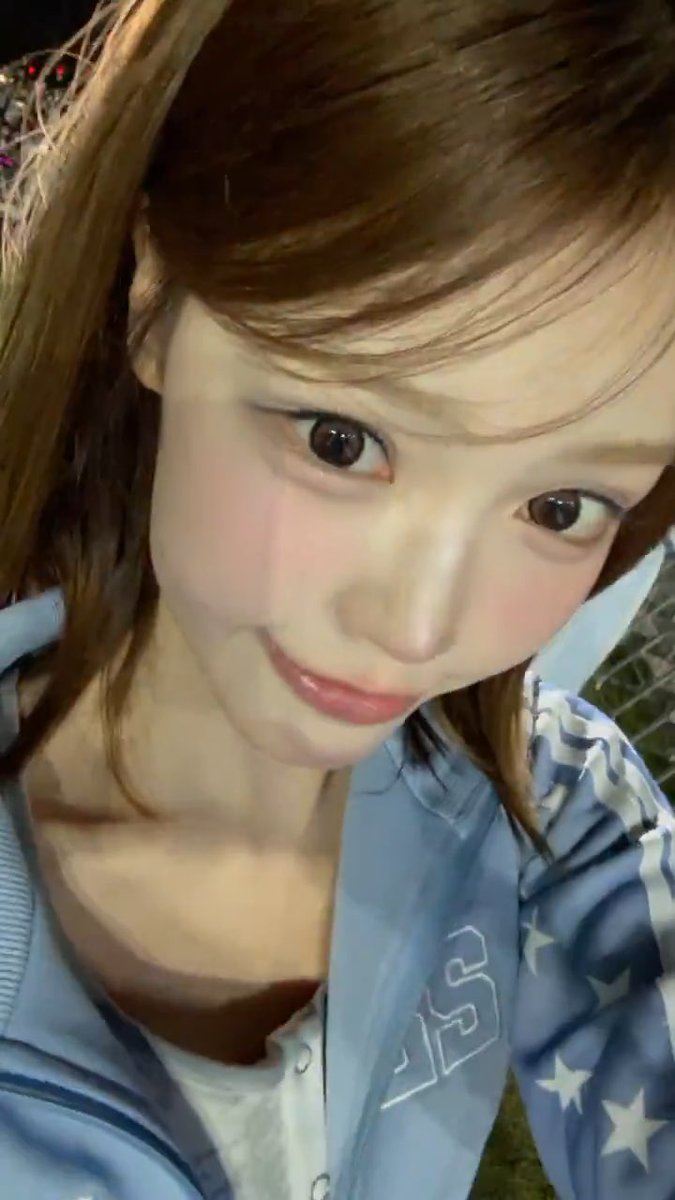A close-up selfie of a young woman with light brown hair and a blue star-patterned jacket over a white shirt, posing with a slight pout in a dimly lit setting.
