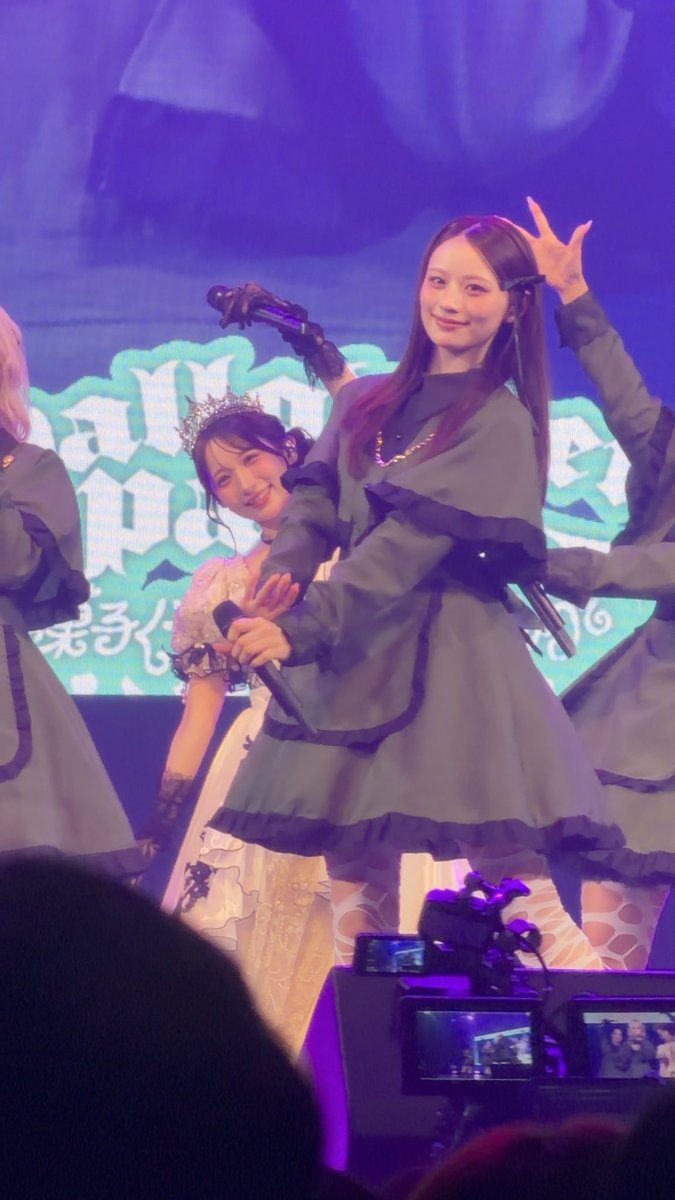 Young female performers in gray gothic-style dresses perform on stage, with one holding a microphone and making a peace sign while another in a tiara poses behind her.