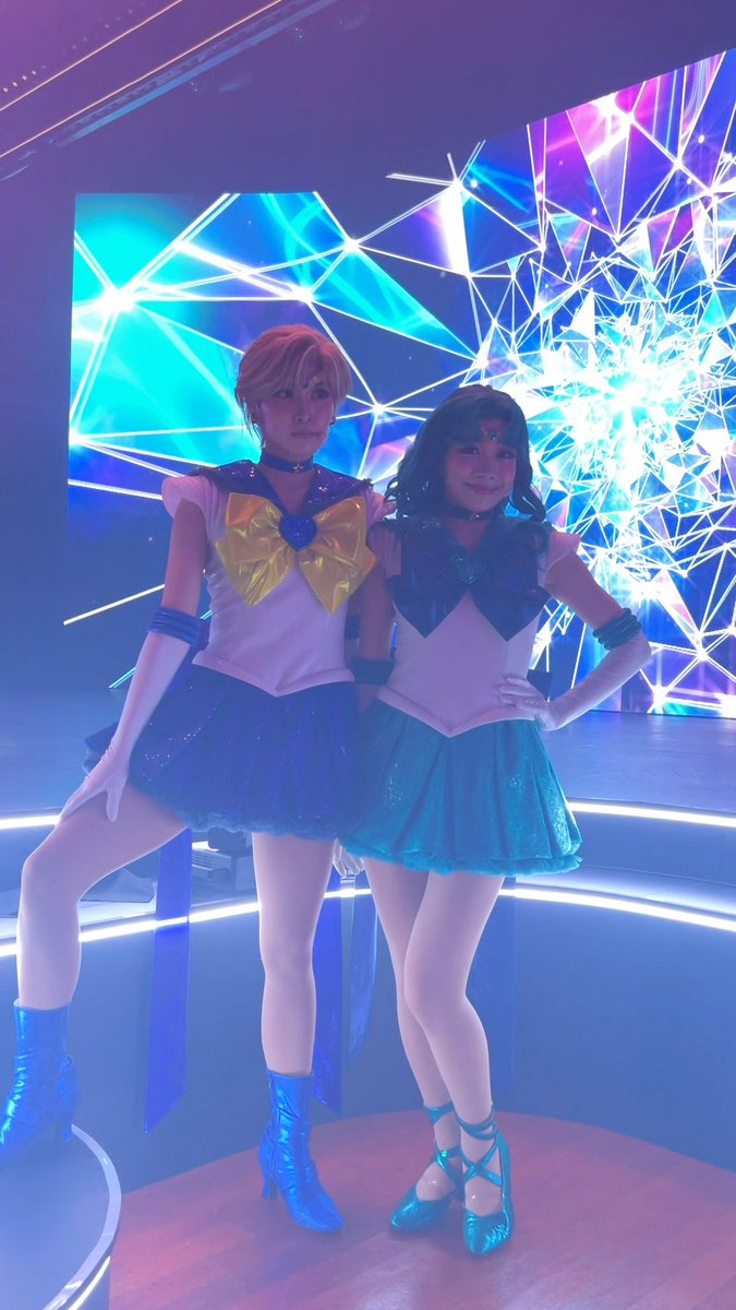 Two women in Sailor Moon costumes posing together in front of a glowing blue and purple digital display.