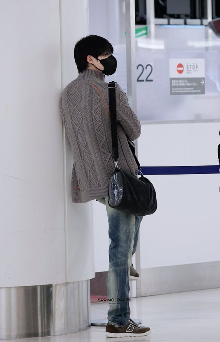 A person wearing a black mask, a gray cable-knit sweater, and jeans stands leaning against a wall while carrying a black shoulder bag in what appears to be an airport setting, with a 'STOP' sign and the number '22' visible nearby.