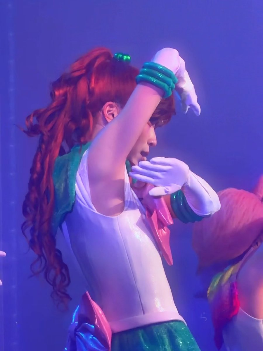 A performer in a Sailor Jupiter cosplay outfit with red hair and green bangle bracelets performs on stage under blue lighting.