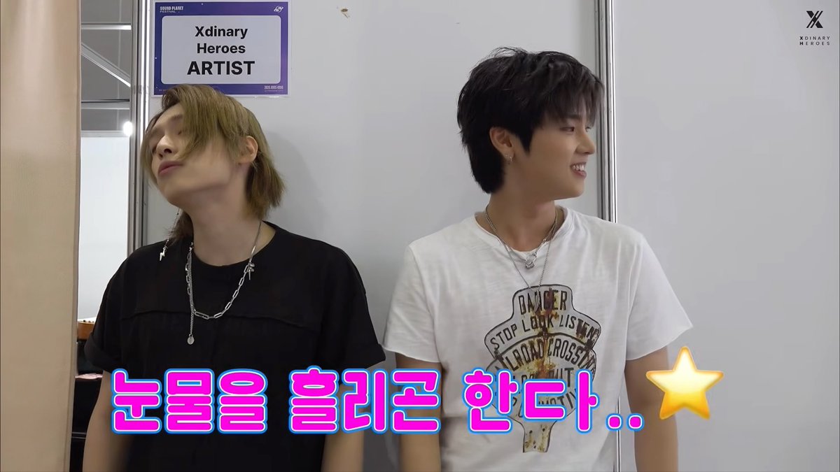 Two young men stand side by side backstage, one looking away shyly while the other smiles broadly, beneath an "Xdinary Heroes" placard labeling them as "ARTIST." The pink Korean text overlay reads "눈물을 흘리곤 한다.." (meaning "tears keep flowing.."), paired with a star emoji, humorously exaggerating the awkward, shy reaction to praise.
