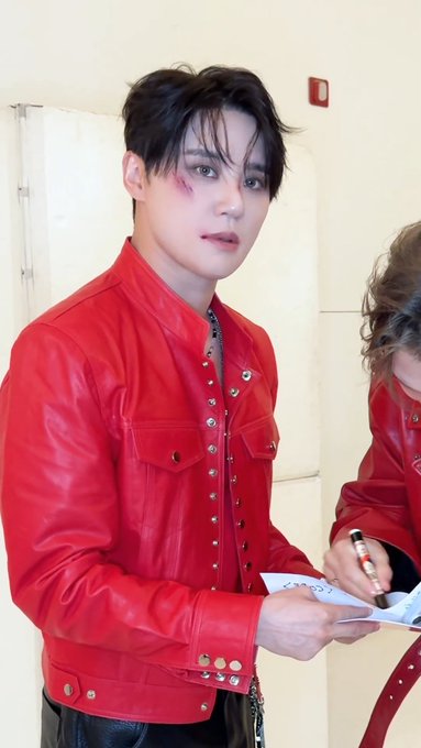A young man with styled dark hair wearing a bright red leather jacket with silver studs, appearing to sign something while another person in red is partially visible.