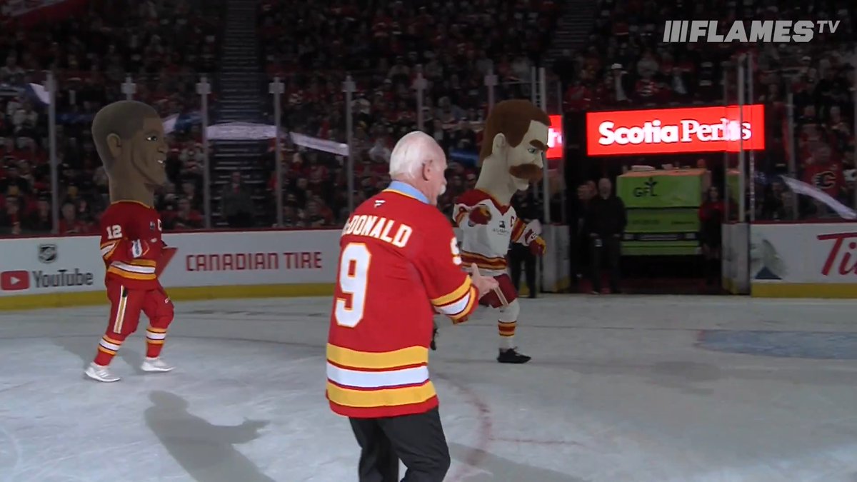 A Calgary Flames hockey player wearing a #9 McDonald jersey stands on the ice between two oversized mascot heads, with the post joking that the bobblehead mascot resembles "Larry the cucumber from Veggie Tales." Visible signage includes "FlamesTV," "Scotia Perks," "Canadian Tire," and "YouTube" in the arena background.