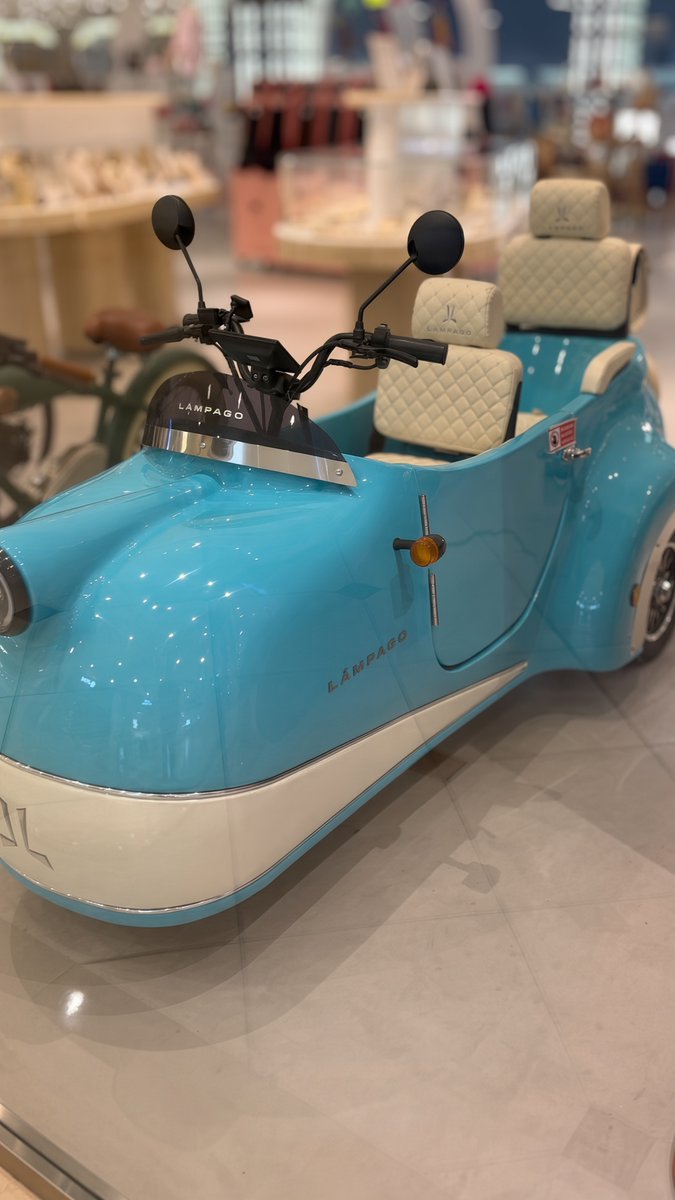 A blue and white three-wheeled electric scooter labeled 'LAMPAGO' on display in a mall setting, featuring quilted cream-colored seats and chrome accents.