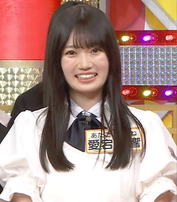 A young Japanese woman with long dark hair, wearing a white blouse and dark bow tie, smiles brightly on a colorful TV studio set featuring bright lights and geometric panels. She wears name tags in Japanese, including one reading "愛宕" (Atago), identifying her as a member of the idol group HKT48 during a variety show appearance.