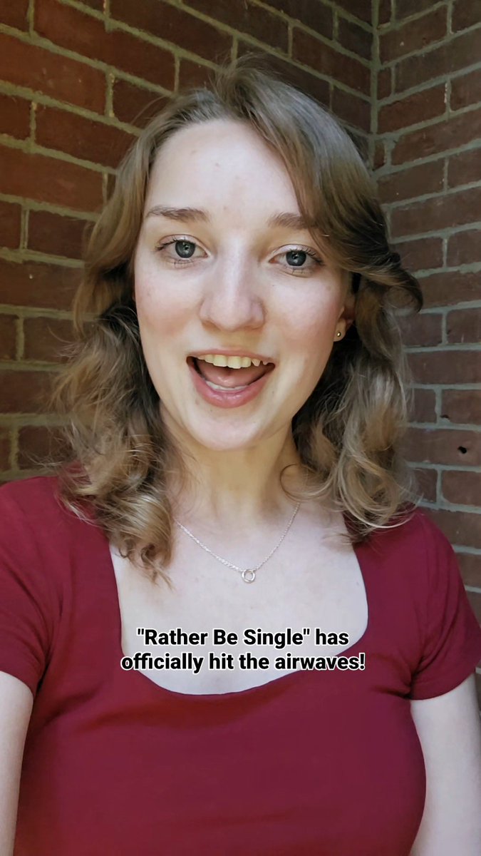 Selfie-style video still of a smiling young woman in a red top against a brick wall, with the caption "'Rather Be Single' has officially hit the airwaves!"