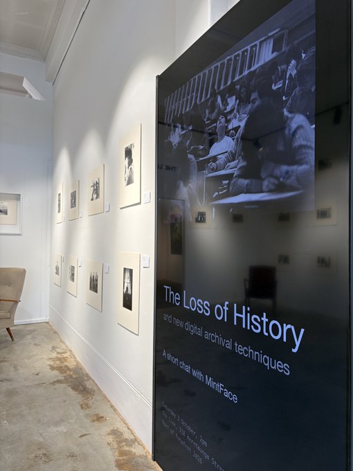 A photo of an art gallery interior featuring a large display titled "The Loss of History" about digital archival techniques, with smaller framed black and white photographs displayed along a white wall.