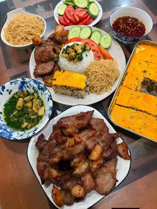 A spread of food includes a large plate with rice, noodles, cucumber, tomato, and meat, accompanied by a separate dish of braised pork, a yellow baked dish in a pan, and small side bowls of sauce and greens.