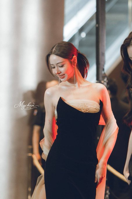 A woman in a black and white dress smiles warmly, bathed in red and orange light at an event, with a 'Mychain' watermark visible.