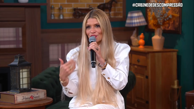 A blonde woman in a white outfit speaks into a microphone during what appears to be a televised segment for "#CABINEDEDESCOMPRESSÃO" (visible in the upper right), seated in a cozy studio setting with a "Home Sweet" book and decorative lamp nearby. Her animated expression and hand gestures align with the post describing her heated commentary criticizing someone named Maíra for "laughing, inciting people, and coming out looking good in photos."