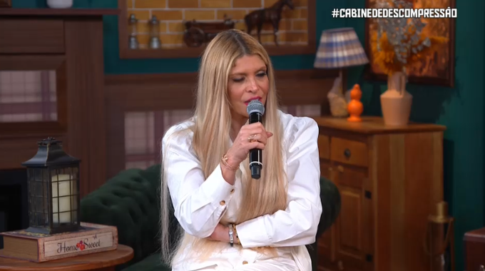 A blonde woman in a white blouse sings into a microphone in a cozy, vintage-styled room with a lamp and horse figurine in the background. The text "#CABINEDEDESCOMPRESSÃO" appears in the top right, matching the post about the singer discussing delivering "fire in the hay" for the audience.