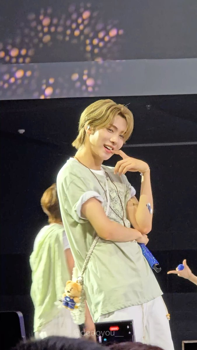 A blonde K-pop idol wearing a light green t-shirt and white pants smiles on stage during a concert, with another member visible behind him and an audience member filming with a phone.