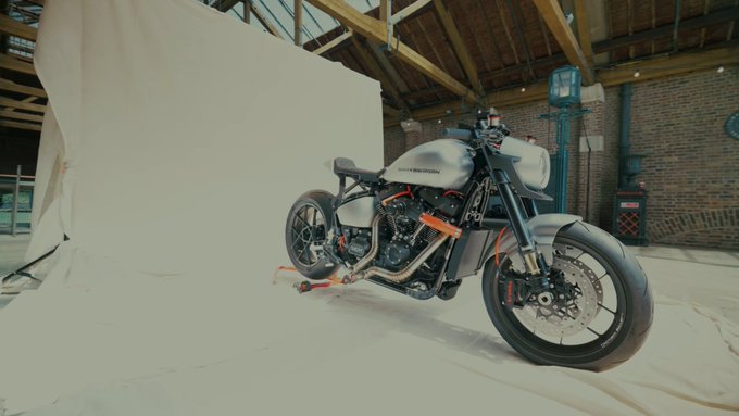 A custom grey Harley-Davidson motorcycle with orange accents is displayed on a white backdrop in a warehouse-like space.