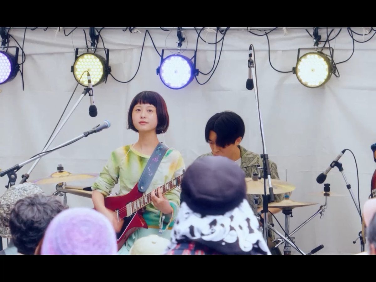 A female musician with a bob haircut plays a red electric guitar on stage under stage lights, with a drummer behind her and audience members visible in the foreground.