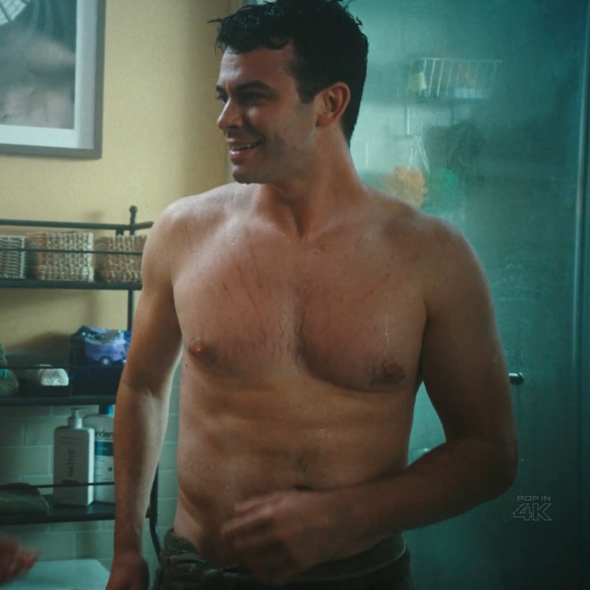A shirtless man stands in a bathroom, smiling slightly as he looks to the side, with visible scratches on his chest and a soft midsection that contrasts with his defined arms. The "POP IN 4K" watermark appears in the bottom right corner, echoing the post's playful question about how someone can have defined abs while still having a belly.