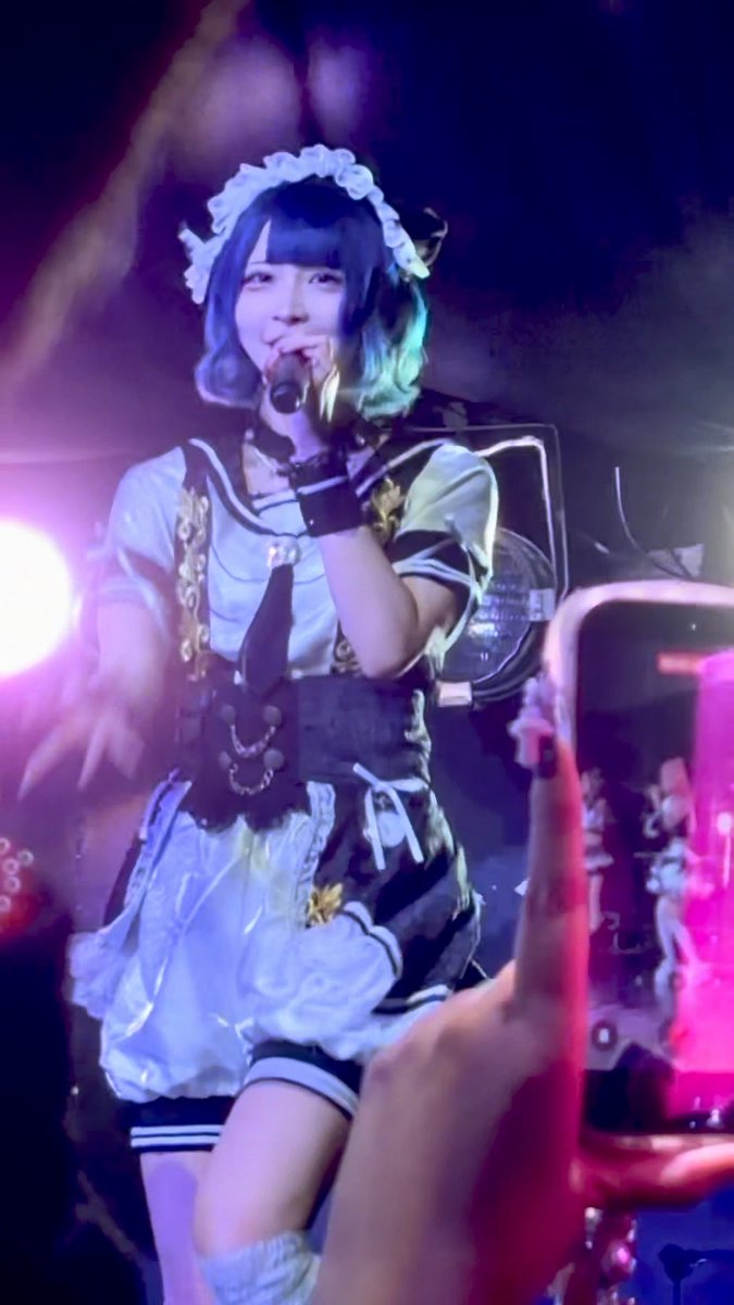A concert photo of a Japanese idol performing on stage, wearing a blue and white maid-style outfit with black gloves, singing into a microphone under purple stage lighting.