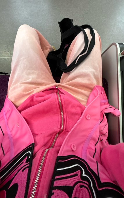 A close-up photo of a person sitting in an airplane seat wearing a pink hoodie with black designs and light pink pants with black straps.