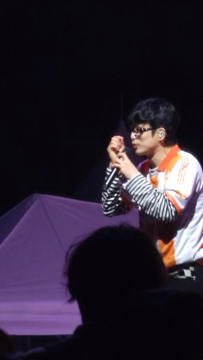 A performer on stage, dressed in a striped shirt and orange and white jacket, sings into a microphone while wearing dark glasses.