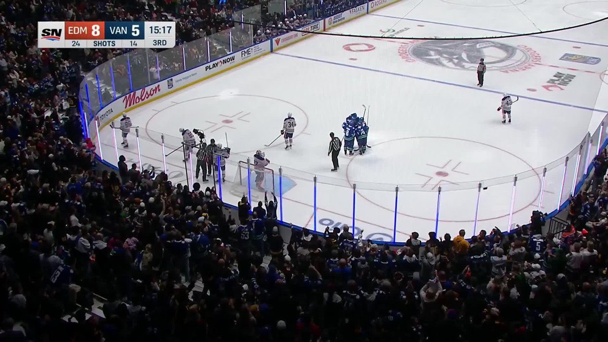A crowd watches an NHL game at Rogers Arena with the scoreboard showing the Edmonton Oilers leading the Vancouver Canucks 8-5 with 15:17 left in the 3rd period and shots 24-14, as the home fans react following a goal that has cut into what was once an 8-2 Oilers lead. Players from both teams and referees gather near the Canucks' net after the play.