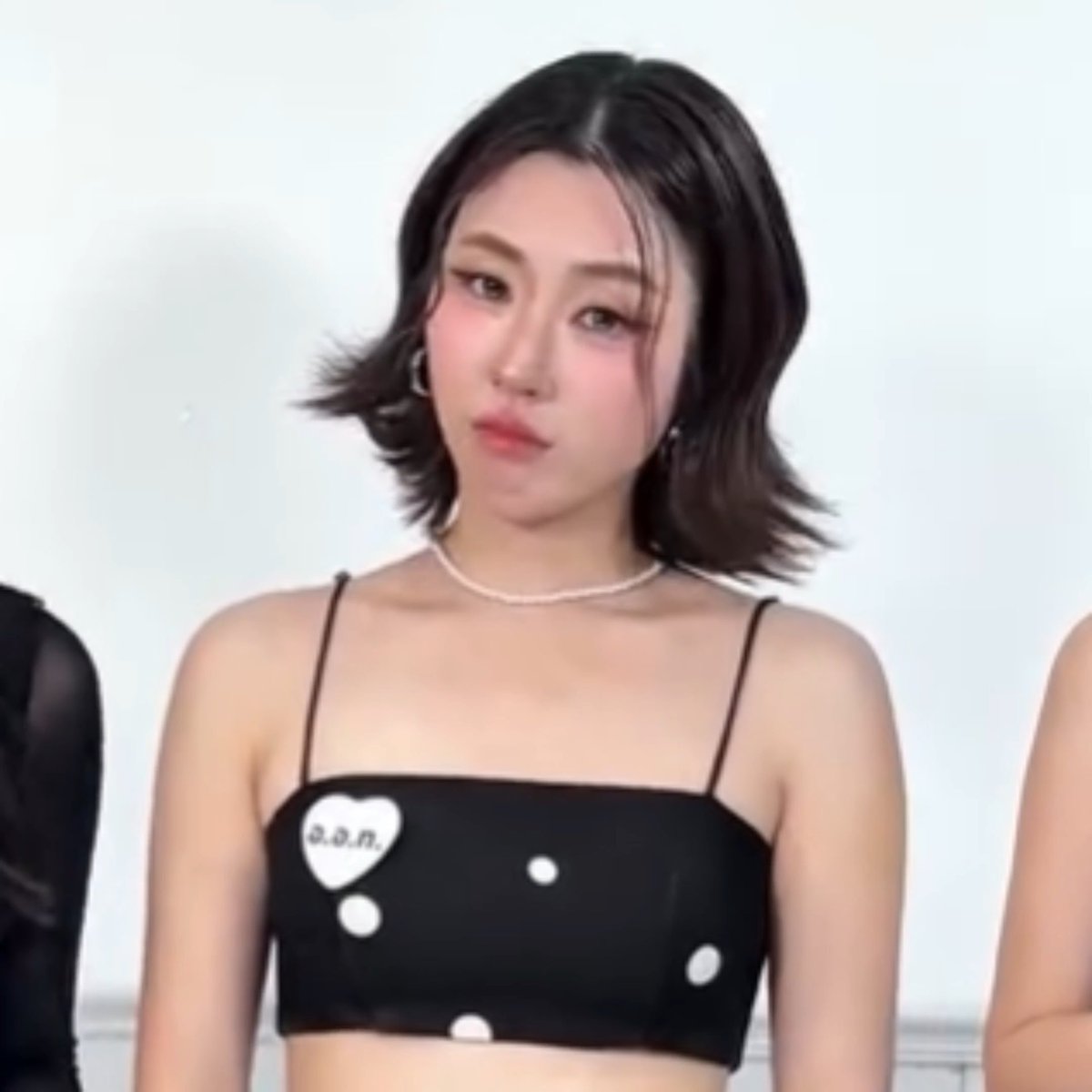 A close-up photo shows a young woman with short dark hair wearing a black crop top with white polka dots and a heart patch reading 'a.o.n.'