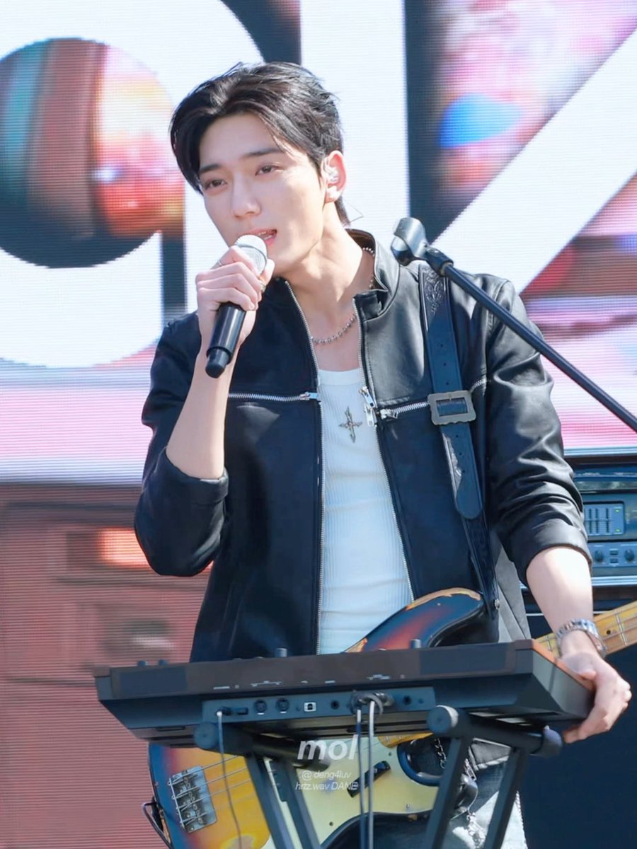 A young male performer in a black leather jacket singing into a microphone while playing a keyboard on stage, with a guitar visible and 'marcel' text.