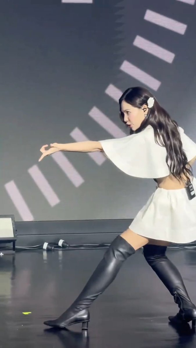 A young woman performs on stage wearing a white cropped top, white skirt, and tall black boots, striking a dance pose against a backdrop with diagonal stripes.