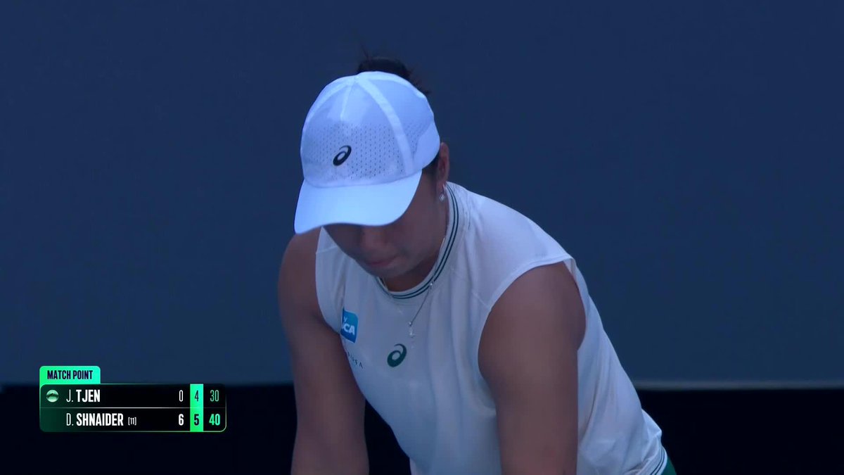 A female tennis player in a white Asics cap and tank top stands with her head bowed against a blue backdrop, as the scoreboard shows "MATCH POINT" with D. Shnaider leading J. Tjen 6-5 (40-30) in their Beijing China Open match.