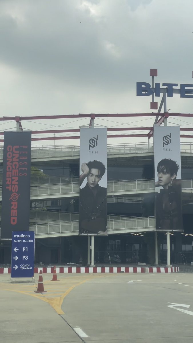 Large vertical banners promoting the 'PERSE'S UNCENSORED CONCERT' featuring portraits of two male artists hang from a stadium structure, with a blue directional sign in the foreground pointing toward parking and a coach terminal.