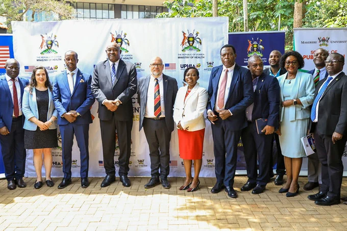 Chargé d'Affaires Cleverley poses with about 20 U.S. and Ugandan officials in a Kampala courtyard. Behind them are banners from the U.S. Embassy, the Uganda Ministry of Health, UPHIA, and the Makerere University School of Public Health.