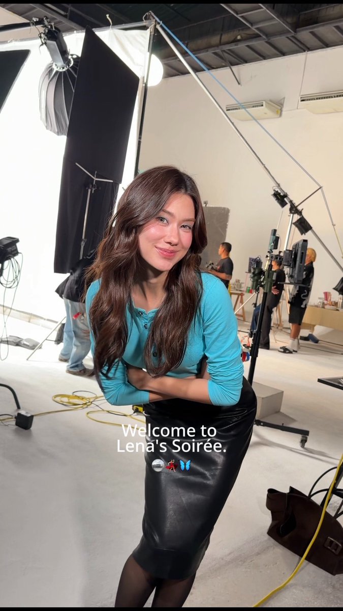 A woman with long wavy brown hair poses confidently in a teal top and black leather skirt inside a photography studio, with lighting equipment and crew visible in the background. The on-screen text reads "Welcome to Lena's Soirée," matching the post's invitation to enter "Lena's Soirée," a promotional collaboration featuring the #SoiréeBag by maisonKEEPS.