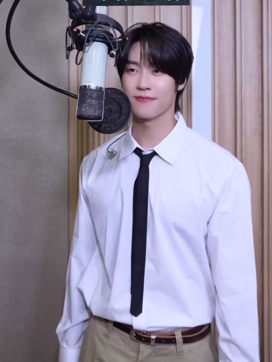 A young man with dark hair, wearing a white shirt and black tie, stands smiling in a recording studio in front of a large microphone.