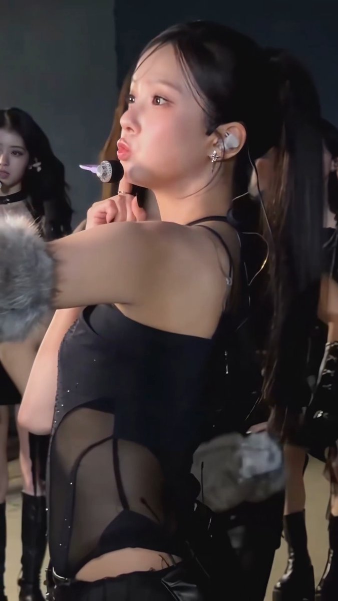 A K-pop performer wearing a black cutout outfit with in-ear monitors and a headset microphone, posing with her hand near her face on stage.