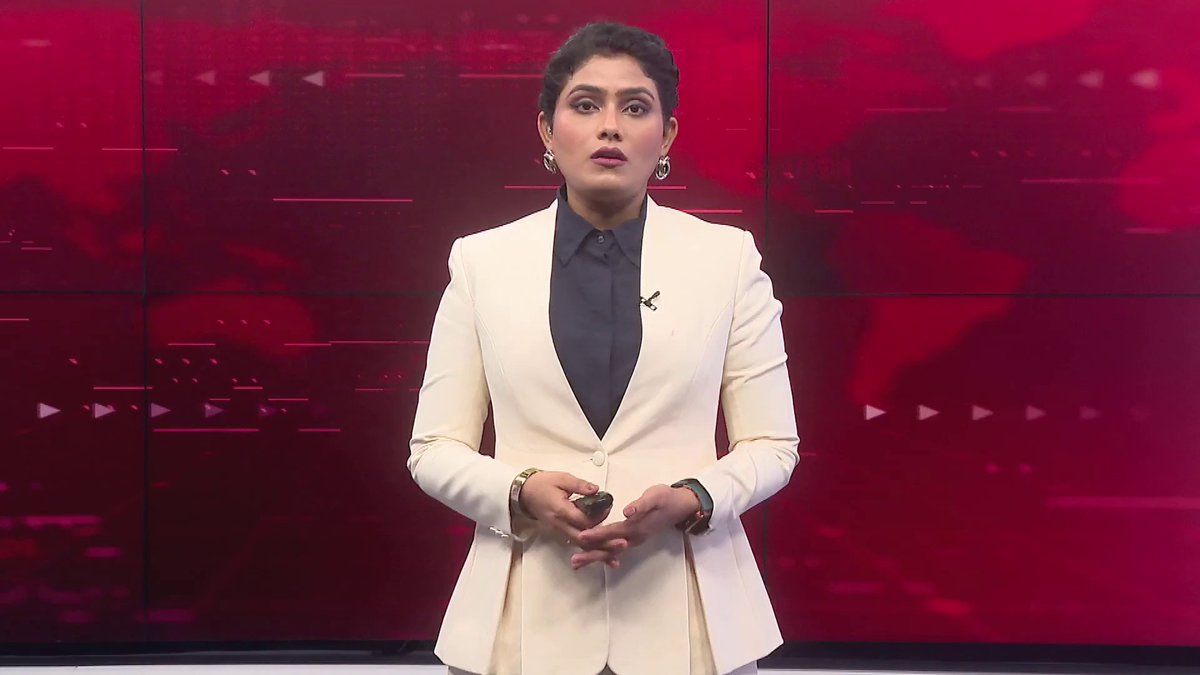 A news anchor in a white blazer and dark blue shirt stands in a studio with a red digital map background, holding a small black device.