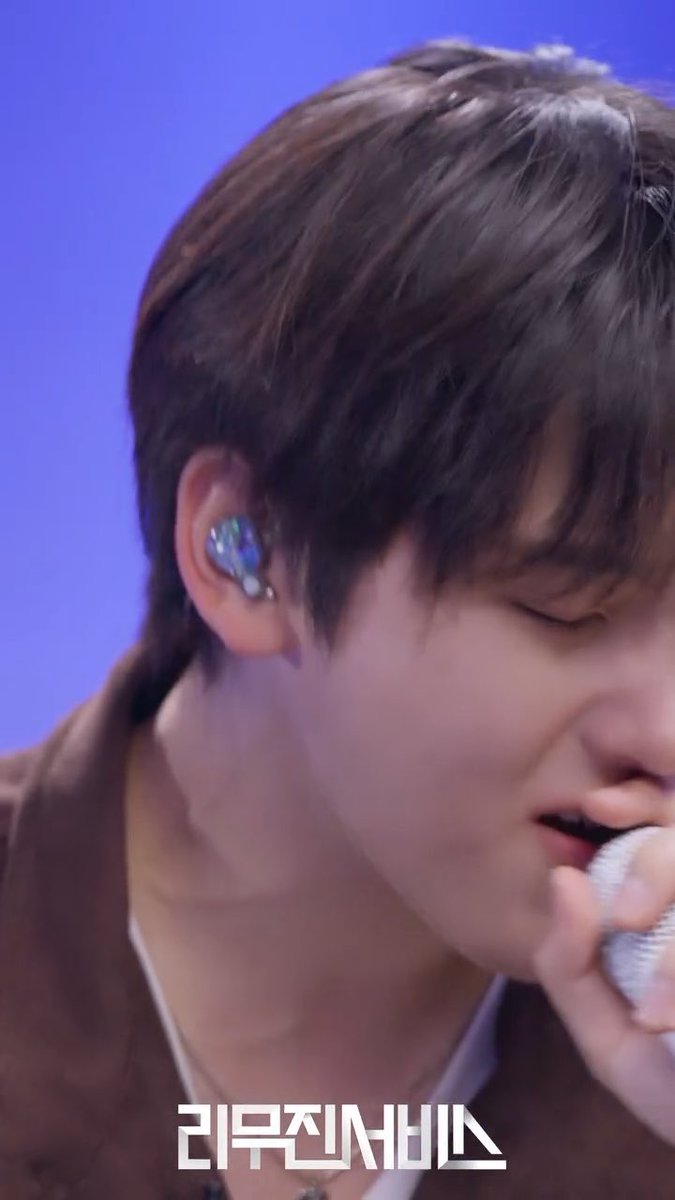 A close-up of a male singer with brown hair wearing an in-ear monitor and singing into a microphone against a purple backdrop, with Korean text '리무진서비스' (Rimousin Service) at the bottom.