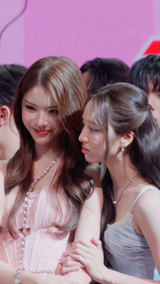Two young women posing closely together at what appears to be an event, one with highlighted brown hair in a pink dress and the other with dark hair in a light blue top, both wearing jewelry.