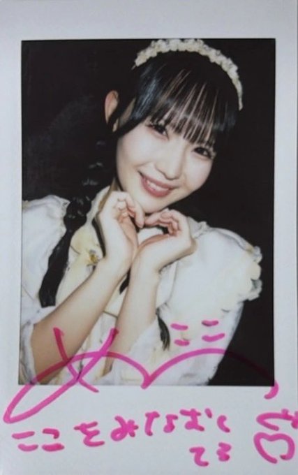 A polaroid-style photo of a smiling young woman making a heart gesture with her hands, signed with pink marker.
