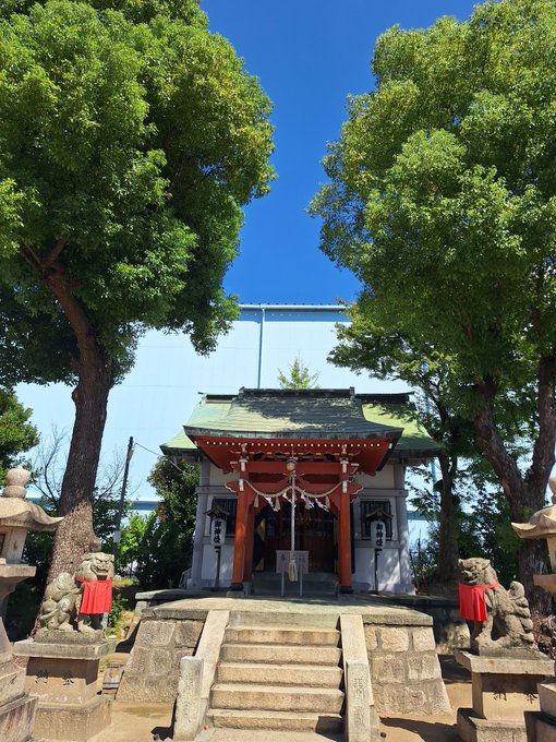 A traditional Japanese Shinto shrine with red and white structures, stone steps, and komainu (lion-dog) statues flanking the entrance, set against a clear blue sky.