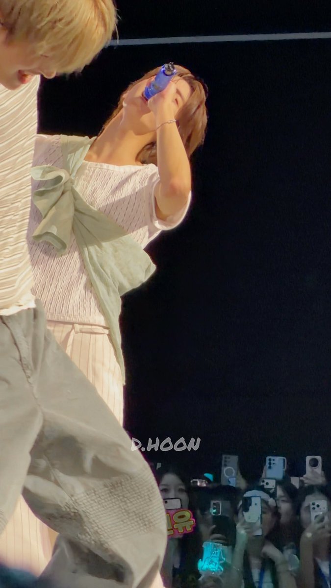 A performer on stage holds a blue water bottle while singing, with another performer visible to the left and an audience filming with phones in the foreground; the watermark "D.HOON" is visible.