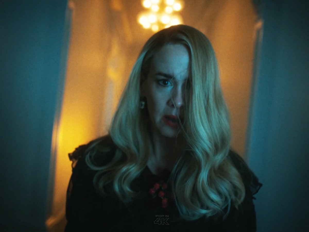 A woman with long blonde hair stands in a dimly lit hallway with a worried expression, wearing a black top with red details, under a glowing chandelier. The "POP IN 4K" watermark is visible at the bottom. The image relates to the post discussing Cordelia from American Horror Story, highlighting her apparent vulnerability in a situation where, as the Supreme, she should have been more formidable.
