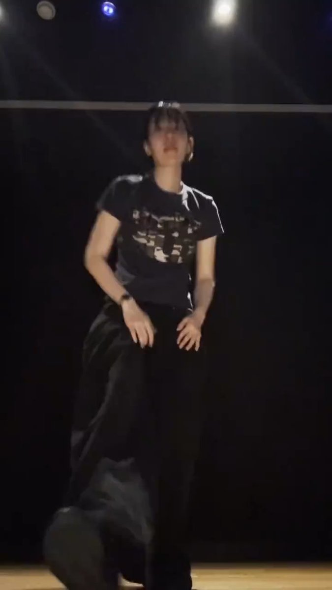 A person on a dark stage wearing a black t-shirt with a graphic design and black pants, captured mid-movement during a performance.