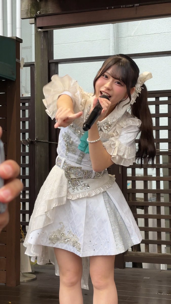 A young woman in an ornate, frilly white idol dress with a jeweled belt sings into a microphone while pointing toward the camera.