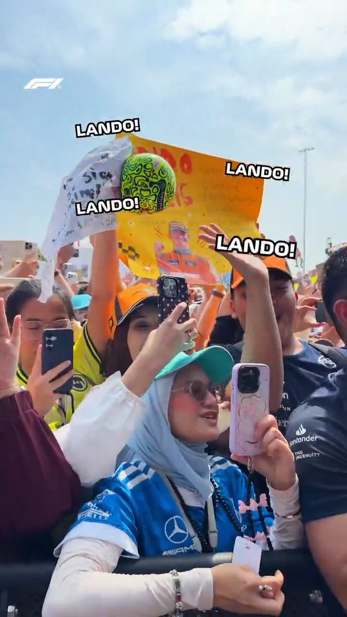 Fans enthusiastically chant "LANDO!" while holding up handmade signs, one featuring a green wrestling mask drawing of Lando Norris, alongside Mercedes and McLaren gear during the BahrainGP fan stage.