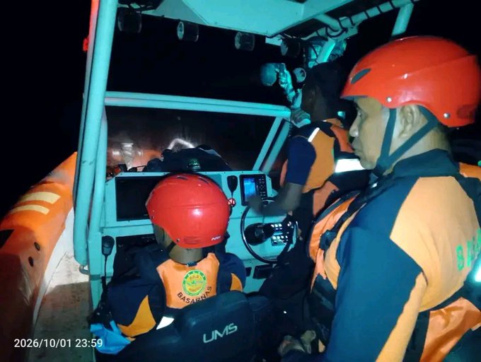 Three people wearing orange life vests and red helmets, marked 'BASARNAS', are on a rescue boat at night, with the timestamp '2026/10/01 23:59' visible on the image.