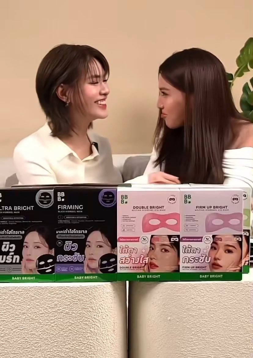 Two young women smiling at each other in what appears to be a product promotion, with Baby Bright skincare masks (Ultra Bright, Firming, Double Bright, Firm Up Bright) displayed in packaging below them.