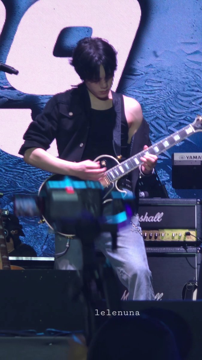 A musician performs on stage playing an electric guitar, wearing a black sleeveless jacket and light jeans, with a watermark "1lelenuna" visible at the bottom.