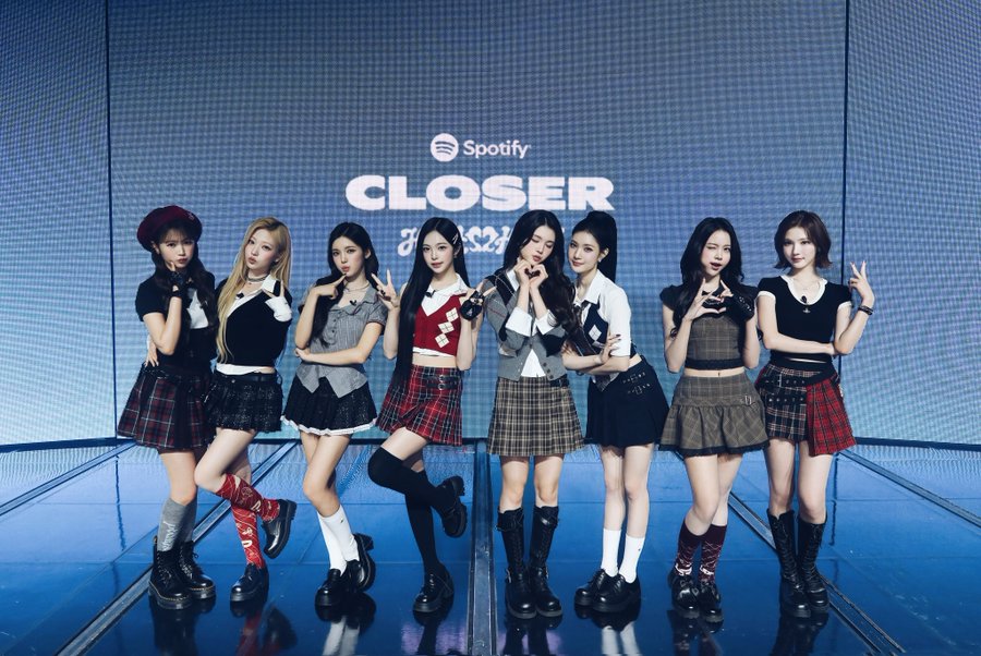 Eight members of the K-pop group Hearts2Hearts stand on a blue stage in schoolgirl-style outfits, posing in front of a screen displaying 'Spotify CLOSER'.